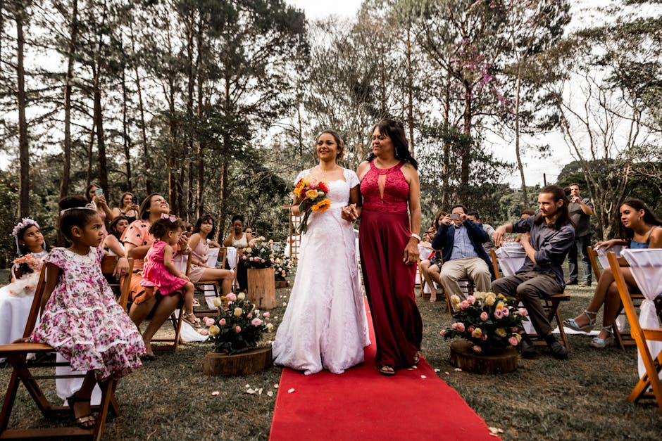 A joyful bride walking with her mother on a red carpet at an outdoor wedding ceremony surrounded by guests