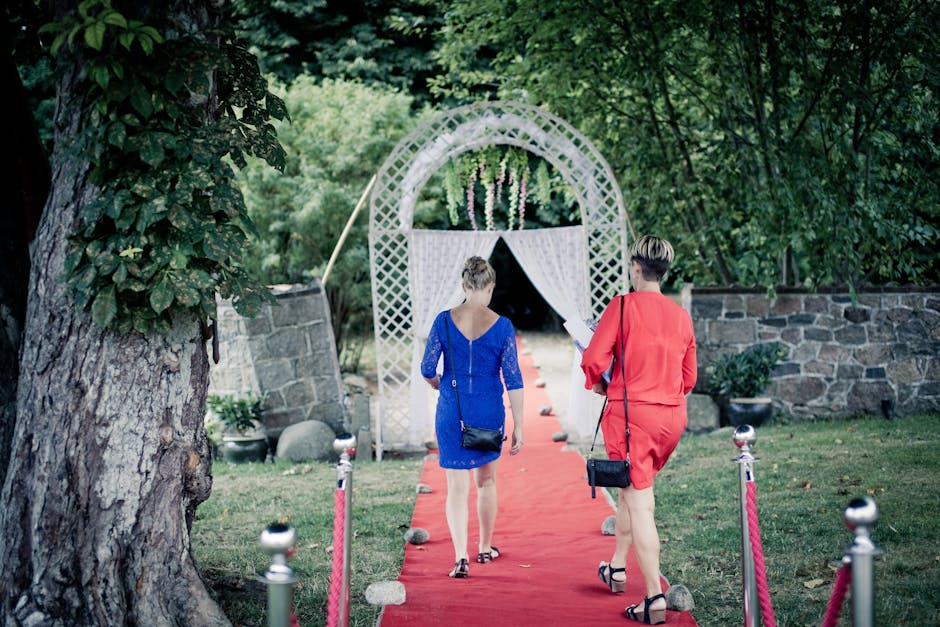 Two women walk on a red carpet through a garden towards a decorative archway
